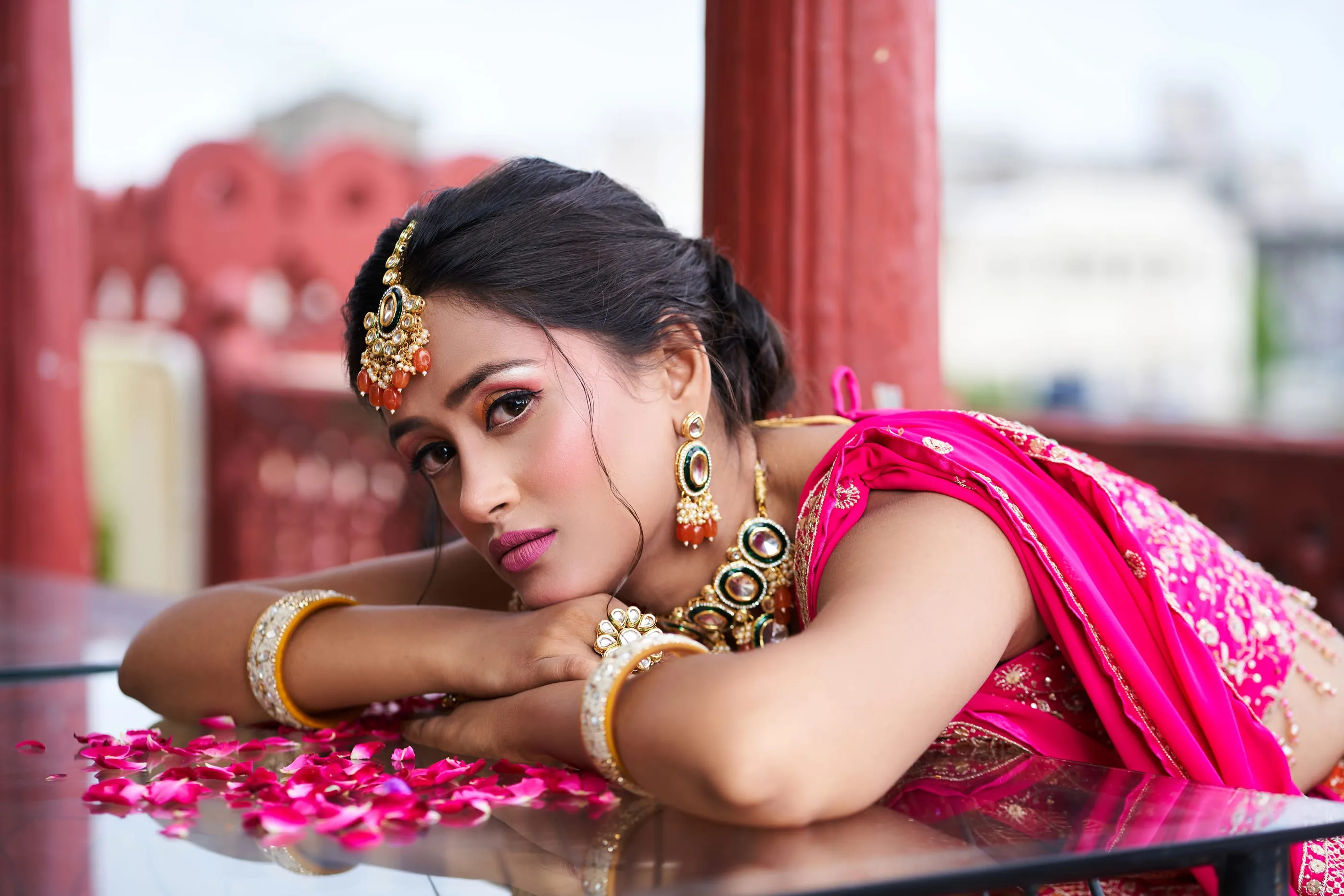 Bride in pink resting on a table scattered with rose petals, Jaipur rooftops behind - Raksha Pareek